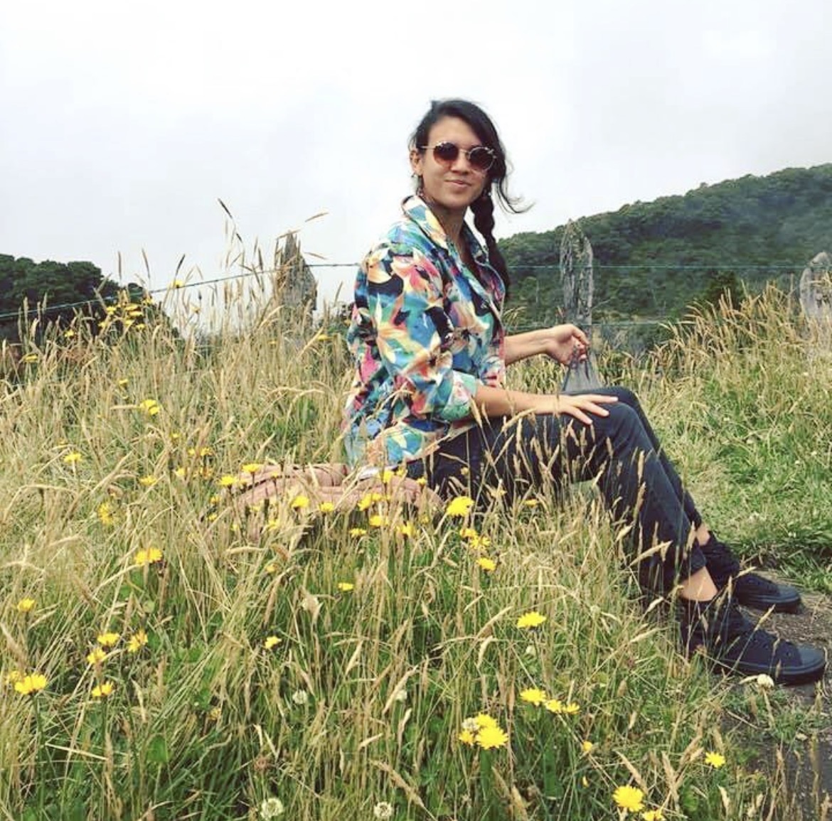 Picture of a woman sitting on a rock on a hill. She is wearing sunglasses a colorful jacket of pink and blue patches, and black pants and shoes. She is surrounded by tall grass and wild yellow flowers.